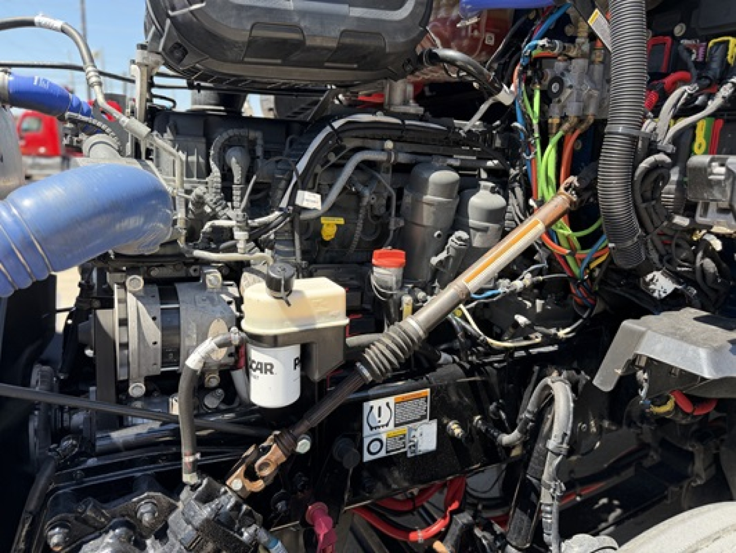 Kenworth truck engine bay with various hoses and wiring; visible reservoir labeled 'COOL' or 'A/C' unclear; multiple colored wires and connectors; black engine components and light tan reservoir caps.
