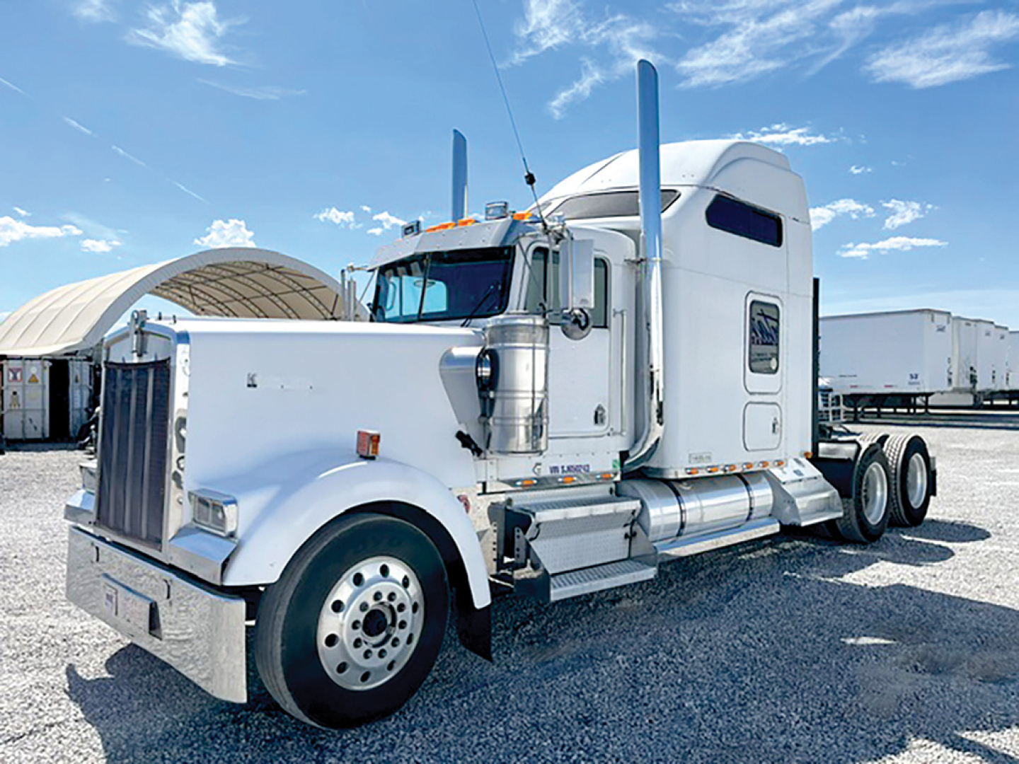 Kenworth W900L tractor with white paint, sleeper cab, chrome exhaust stacks, front bumper, and dual rear axles in a gravel lot.