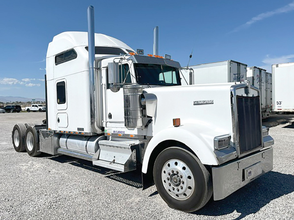 Kenworth W900L tractor with sleeper cab in white, front grille, dual exhaust stacks, large chrome bumper and wheels; appears in a lot with trailers in background.