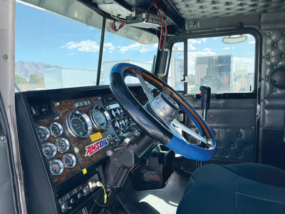 Kenworth W900L truck interior showing dashboard with multiple gauges, steering wheel with blue cover, driver seat, and window view outside.