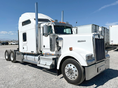 Kenworth W900L tractor with sleeper cab in white, front grille, dual exhaust stacks, large chrome bumper and wheels; appears in a lot with trailers in background.