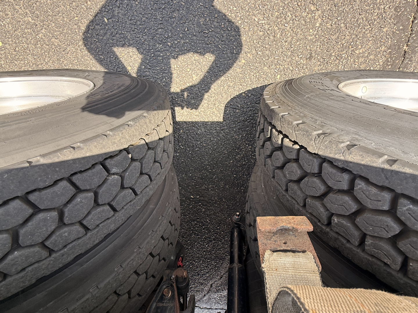 Truck image showing dual rear wheels (tires) on a flatbed truck bed with shadow of a person, rusted heavy hitch area visible between tires.