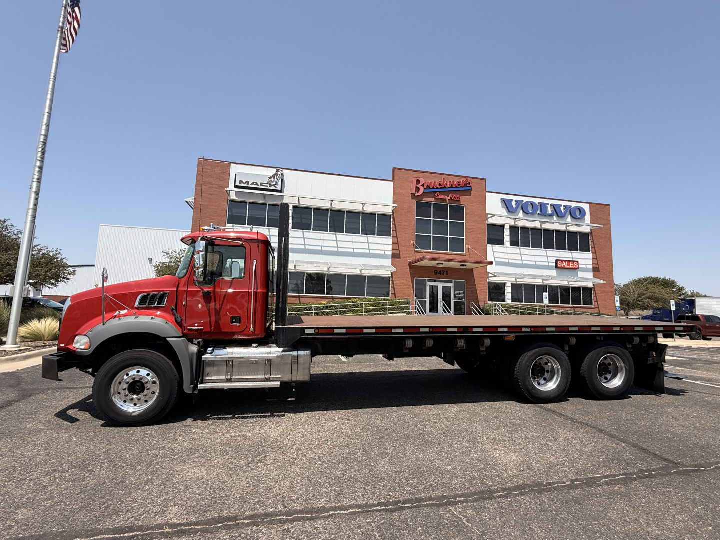 Flatbed truck in foreground with red Mack Granite cab and long flatbed, dealership building in background
