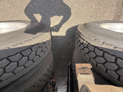 Truck image showing dual rear wheels (tires) on a flatbed truck bed with shadow of a person, rusted heavy hitch area visible between tires.