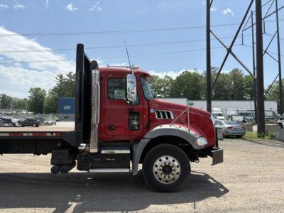Truck side profile showing a red Mack Granite cab with flatbed, black vertical ramps at rear, aluminum wheels, and dealership lot in background.