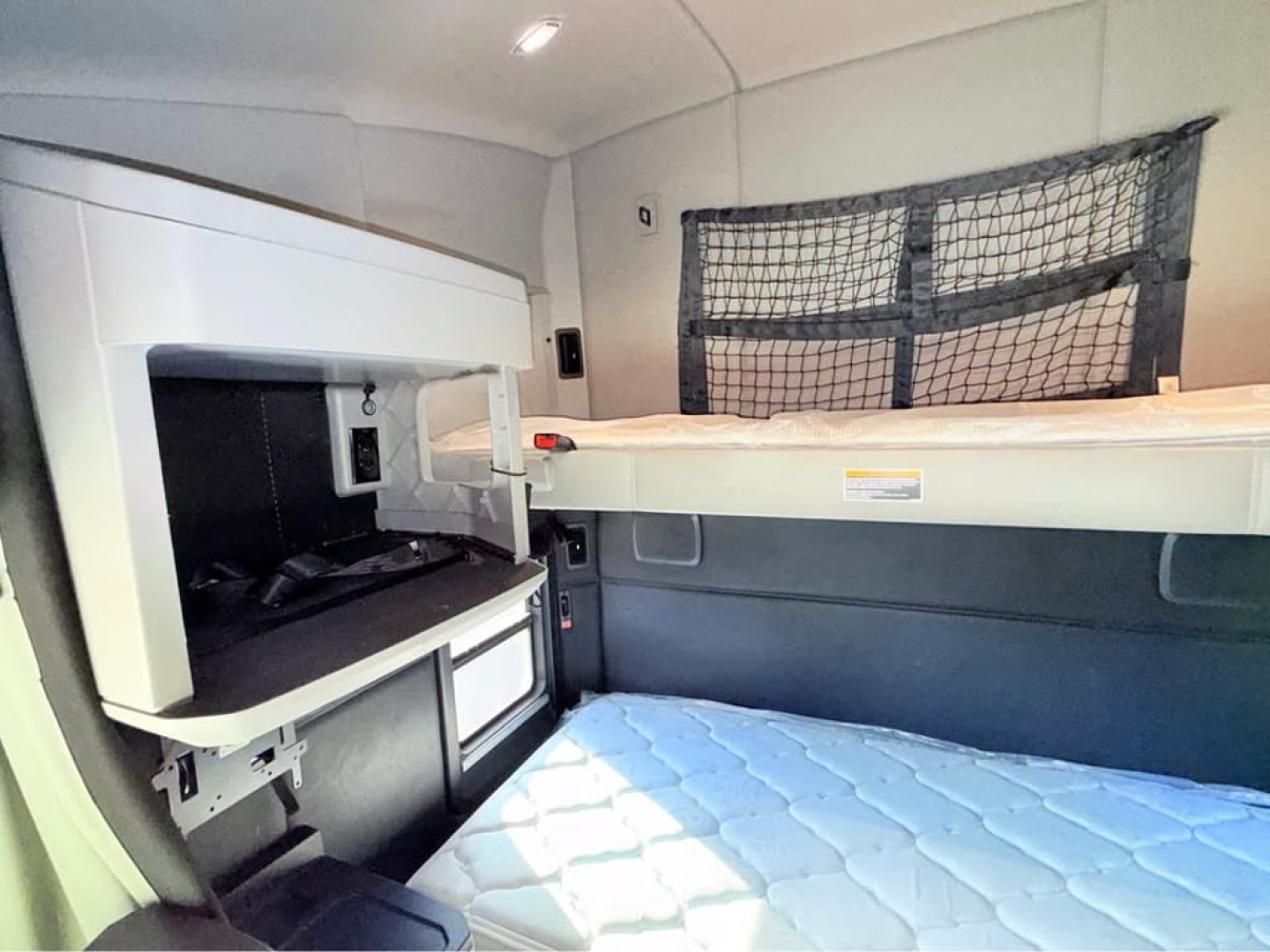 Kenworth T680 sleeper cab interior with bunk bed and overhead storage, showing a small cabinet and netted storage behind the bed. Visible text: none legible from image.