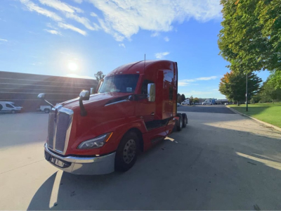 Kenworth T680 semi-truck in red sleeper cab side view showing front grille and headlamp, highway setting with clear sky.
