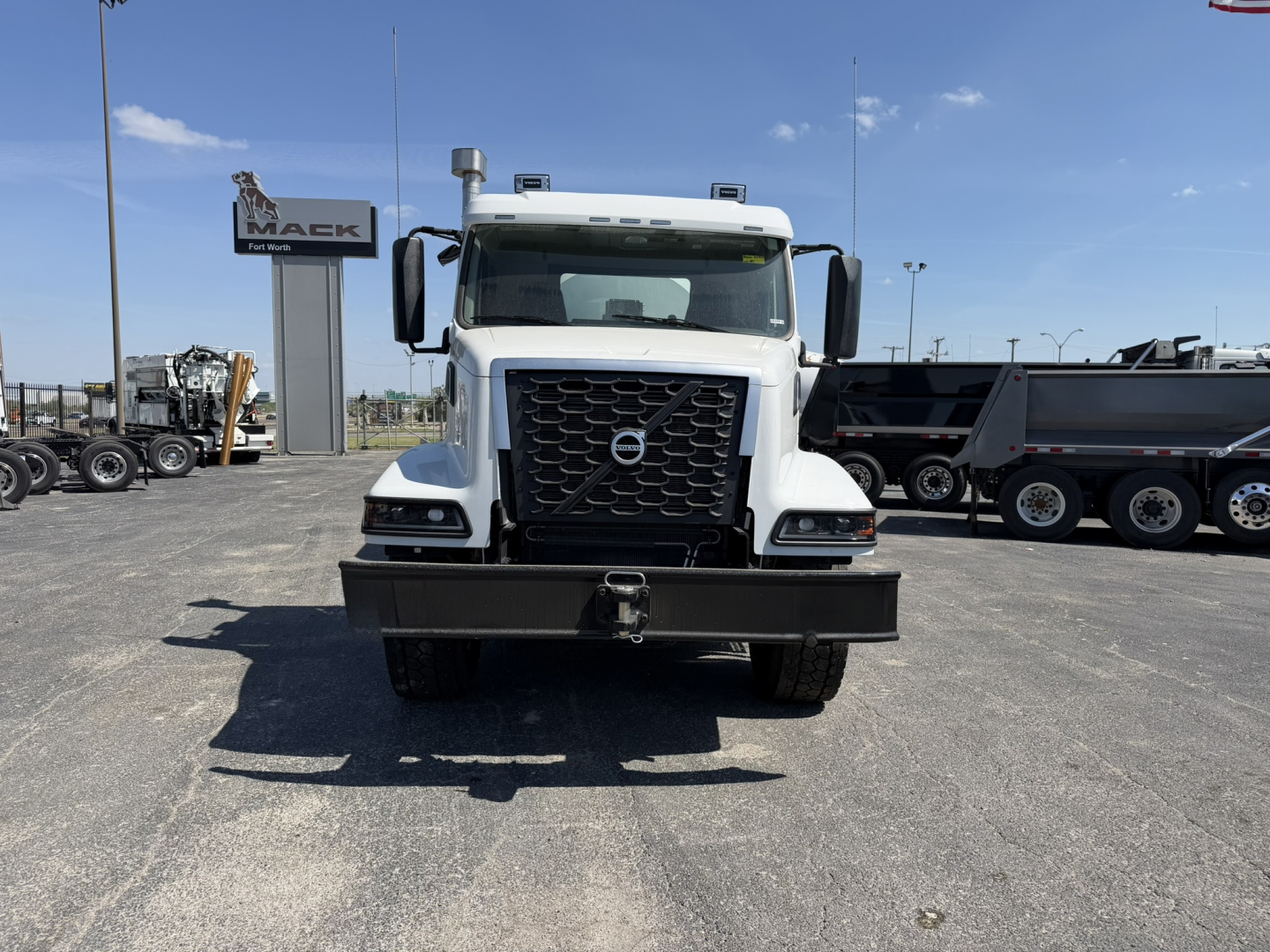 Front view of a white Volvo mixer truck with large black grille, bumper, and towing hook. Volvo emblem on grille; dealership signs in background.
