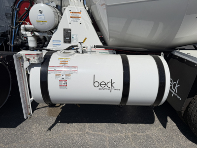 Truck side view showing Beck concrete mixers label on cylindrical tank attached to mixer truck chassis. Visible decals and hoses around the tank with warning labels.