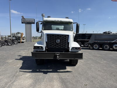 Front view of a white Volvo mixer truck with large black grille, bumper, and towing hook. Volvo emblem on grille; dealership signs in background.
