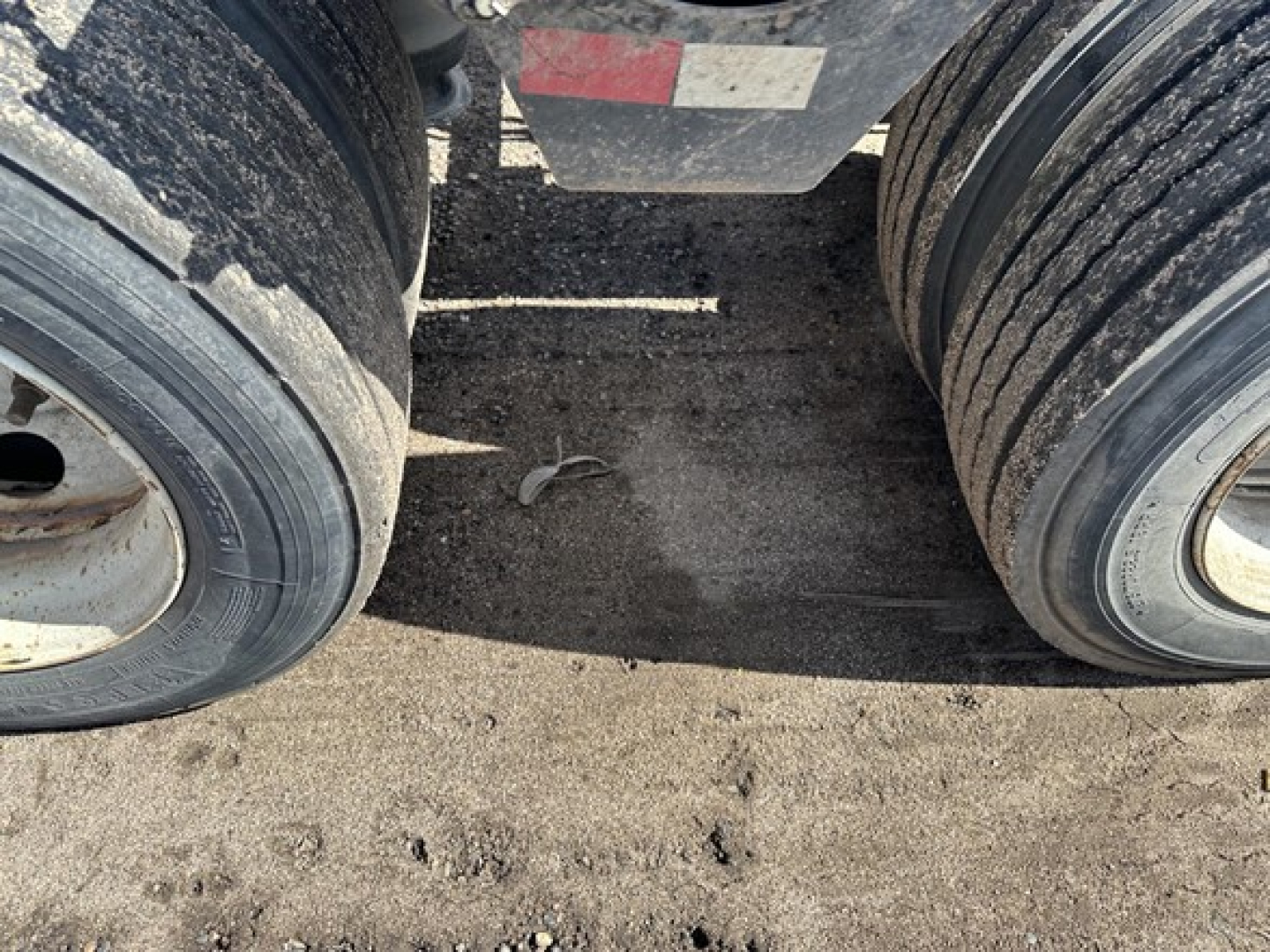 Lowboy trailer axles with dual tires and a dirty deck area between wheels, showing portion of frame and ground surface.