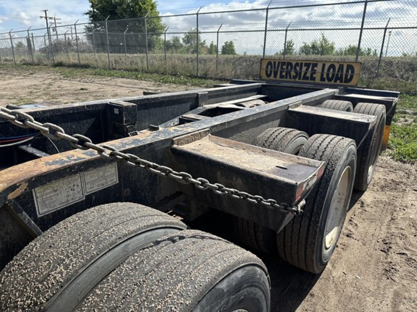 Lowboy trailer pair of axles showing dual tires and chain secured deck with "OVERSIZE LOAD" sign in background.