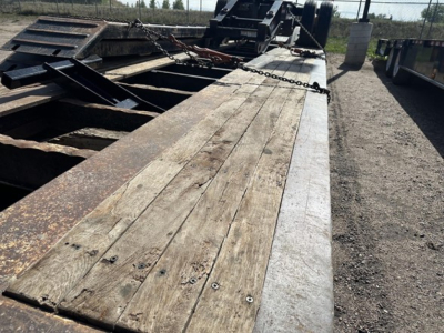 Lowboy trailer deck with weathered wood planks and steel edging; visible chain securing mechanism and heavy equipment hitch in background.
