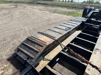 Lowboy trailer deck with metal ramps and chained stake pockets; chain and metal plates along the edge; View shows long flatbed section and dual rear wheels in background.