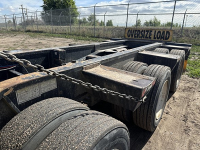 Lowboy trailer pair of axles showing dual tires and chain secured deck with "OVERSIZE LOAD" sign in background.