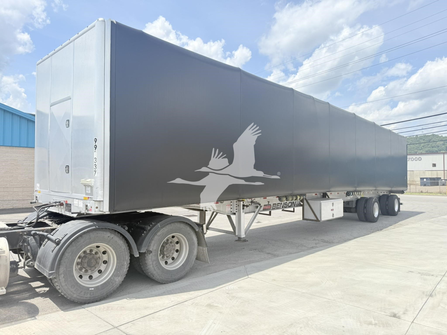Curtainside trailer with a gray exterior and a white logo of two birds in flight on the side; rear doors and undercarriage visible; truck tires in foreground.