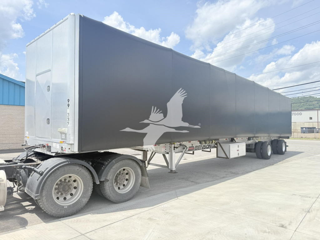 Curtainside trailer with a gray exterior and a white logo of two birds in flight on the side; rear doors and undercarriage visible; truck tires in foreground.