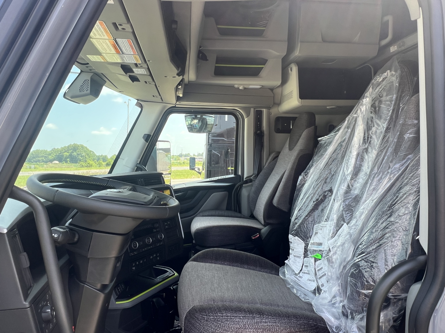 Cab interior of a Volvo VNL truck showing steering wheel, dashboard, driver seat, and passenger seat wrapped in plastic; overhead storage compartments and controls visible.