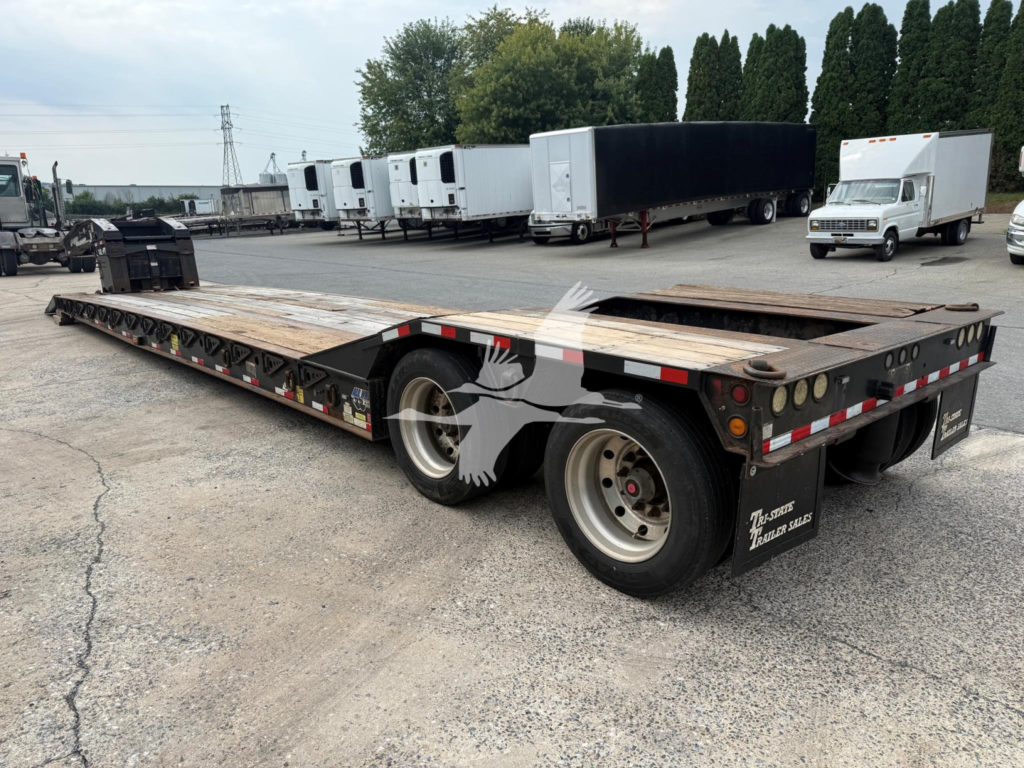 Flatbed trailer with lowboy style, dual rear axles, wooden deck, and black undercarriage. Text on mudflaps reads 'EAGER BEAVER' and other branding; background shows other trailers and a white box truck.