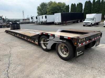 Flatbed trailer with lowboy style, dual rear axles, wooden deck, and black undercarriage. Text on mudflaps reads 'EAGER BEAVER' and other branding; background shows other trailers and a white box truck.