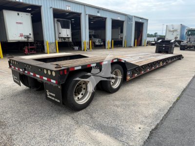 Lowboy trailer showing dual rear axles and long flatbed with wooden deck and wheel wells.