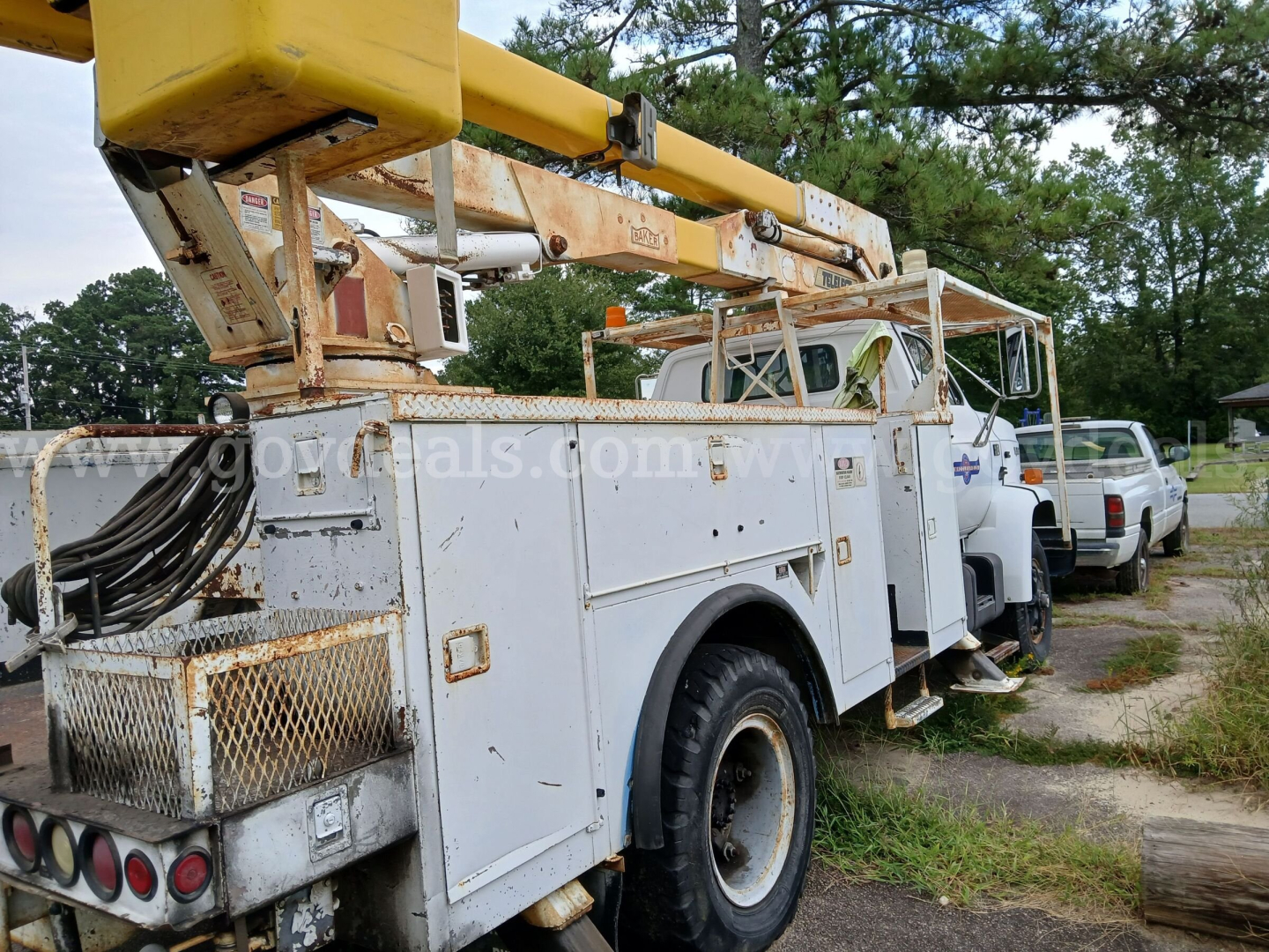 1986 GMC bucket truck with a yellow bucket boom extended over the cab; service body with compartments, dual rear wheels, and utility ladder racks. Visible text includes faint stickers and a license plate area on the side; overall vehicle sh