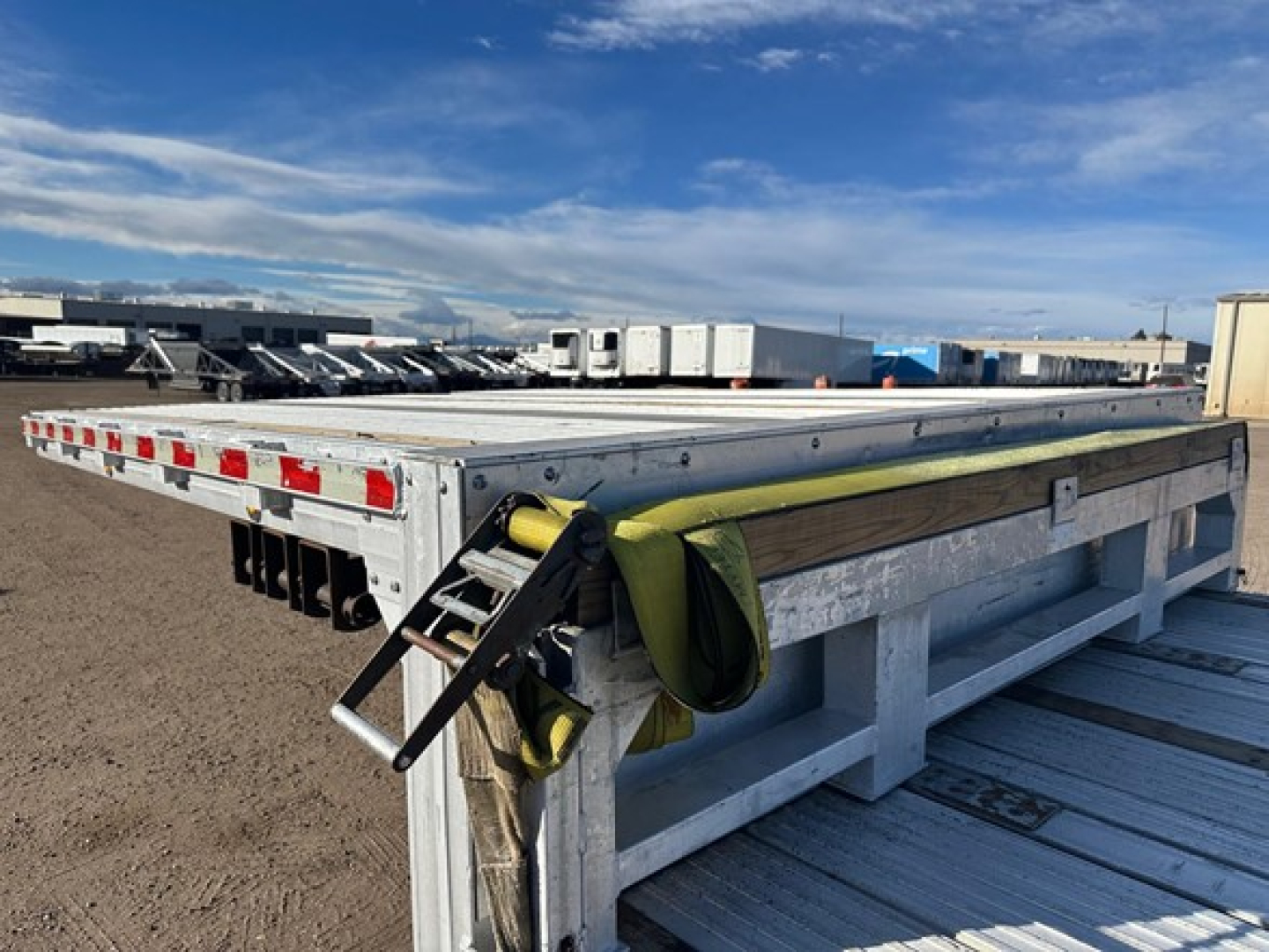 Trailer deck showing aluminum drop deck with reinforced frame, tie-down straps and rear lights along side. Side rails and ramps visible.