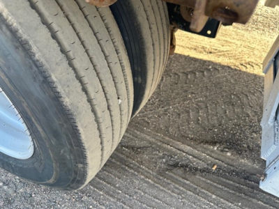 Trailer wheel and dual tire undercarriage view showing aluminum drop deck trailer components and gravel ground.