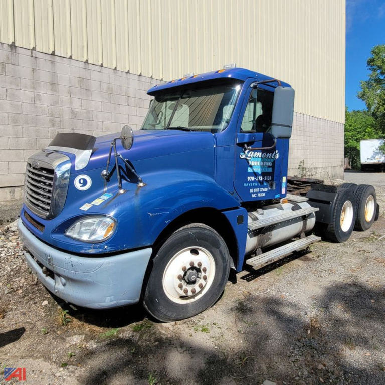Blue Freightliner Columbia 120 daycab front-left view with dual rear axles, engine bay area, and scaffolding on rack; text on door and side reflectors visible.