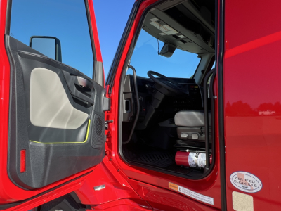 Volvo VNL64T660 truck cab doorway open showing interior seat and steering wheel; door panel with light gray insert and black handle; red exterior; a circular Clean Idle certification sticker on the door frame; blue sky.