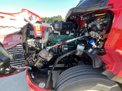 Volvo VNL64T660 engine bay view showing diesel engine and surrounding components in red truck front, with visible hoses, belts, and coolant reservoir.