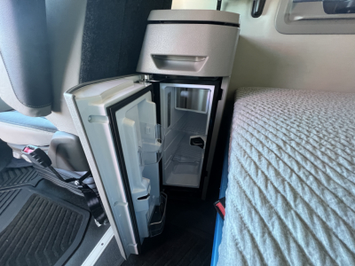 Small fridge or cooler mounted in Volvo VNL64T660 sleeper cab with open door showing interior shelves and a compact freezer section.