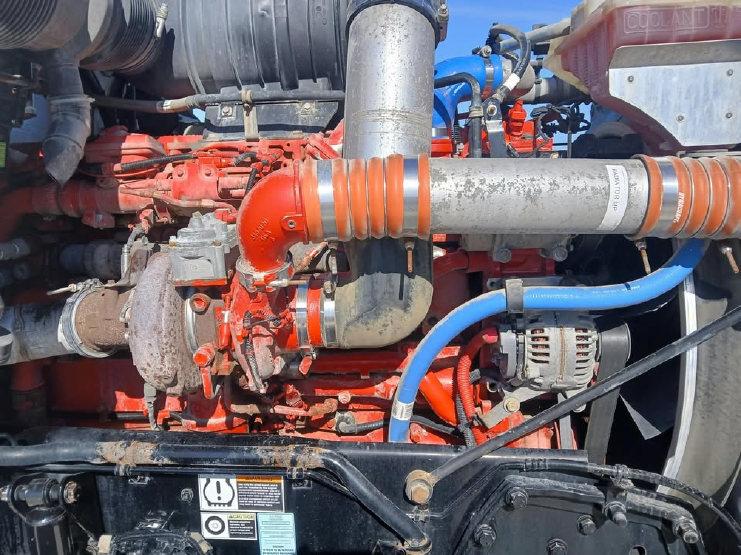 Kenworth T800 engine bay showing red engine block, orange intake manifold, blue hoses, silver exhaust components, and black frame with warning labels.