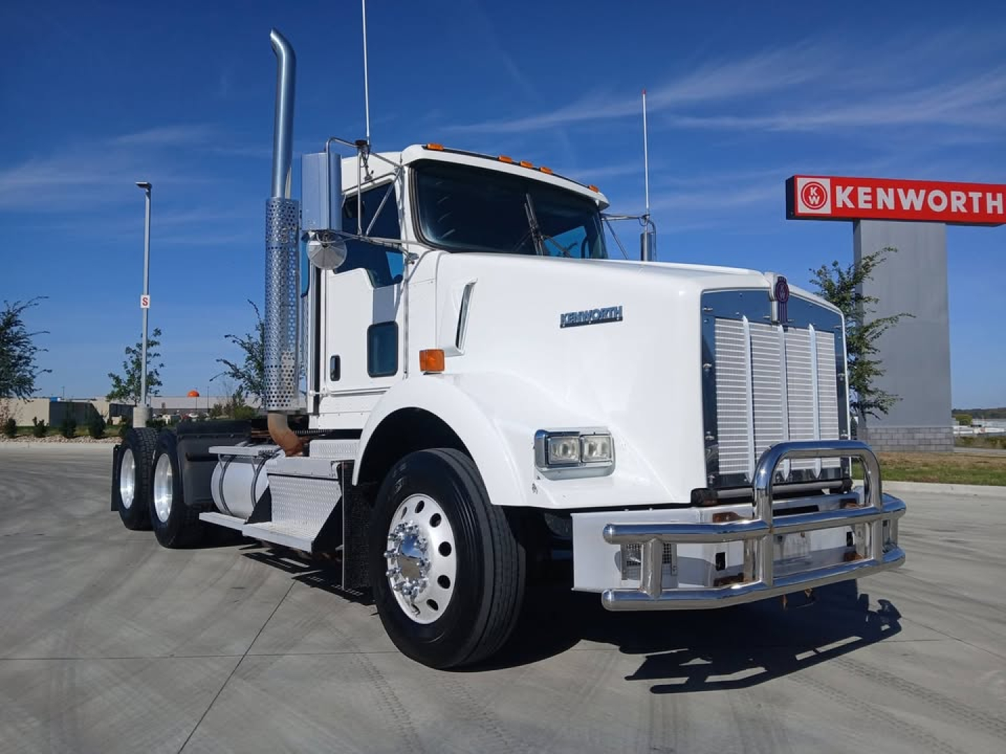Kenworth T800 white conventional sleeper truck showing chrome grille, bumper, vertical exhaust, dual rear axles, and Kenworth sign in background.