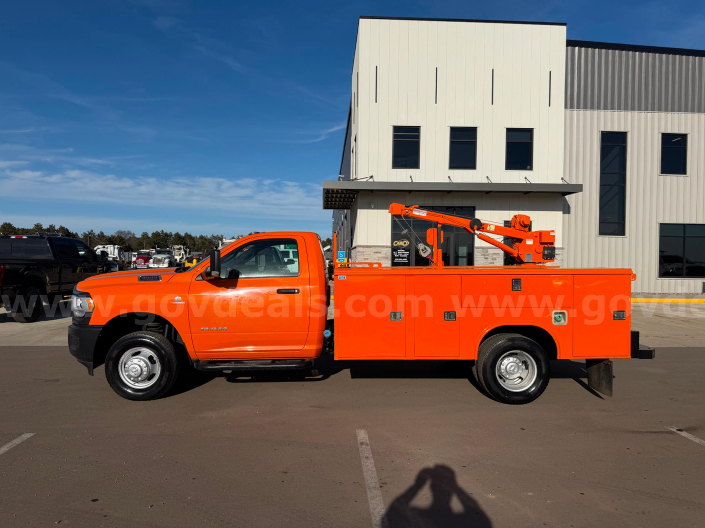 Orange Ram 3500 utility truck with cherry picker in a parking lot, side view showing service body with multiple compartments and a hydraulic boom.
