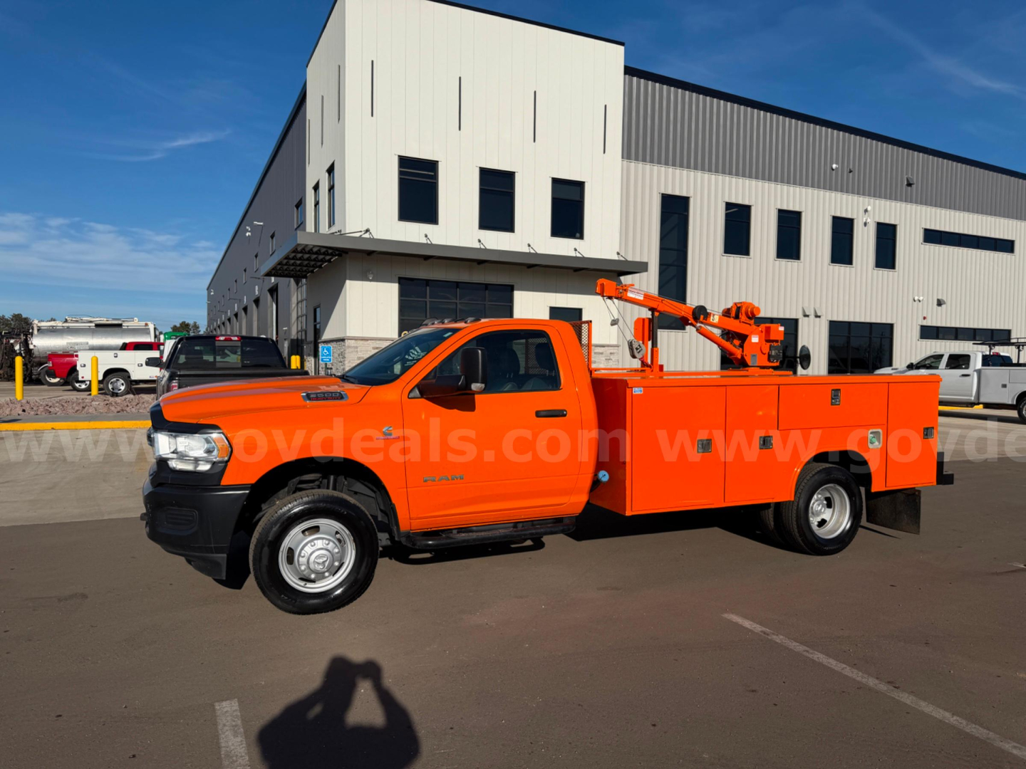 Utility/service truck with orange body and bucket lift, Ram 3500 pickup, side view showing utility compartments and boom arm, dealership lot in background.
