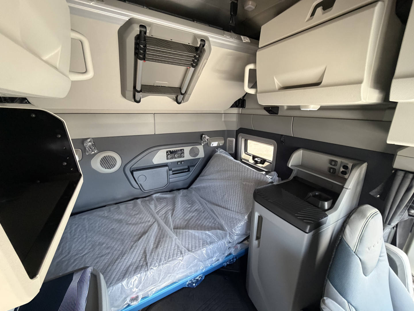 Volvo VNL sleeper cab interior showing bunk bed with plastic cover, overhead storage, cabinets, and control panel near window.