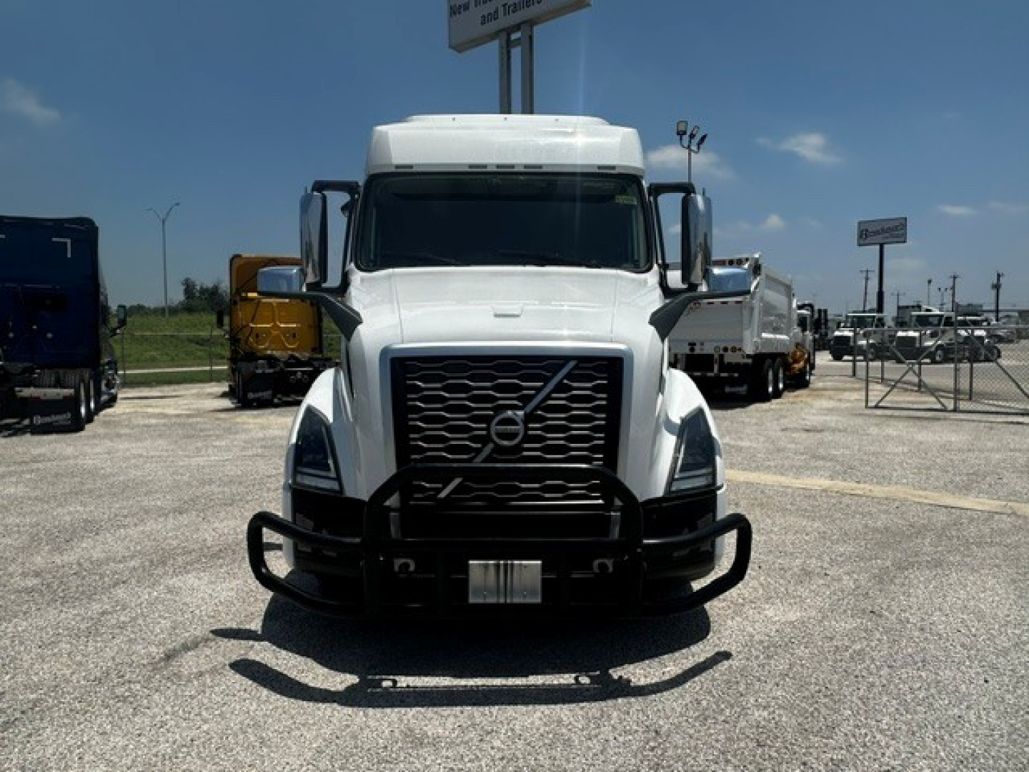 Volvo VNL truck front view with grille guard in a dealership lot. No visible text on the truck, distant signs present but unreadable.