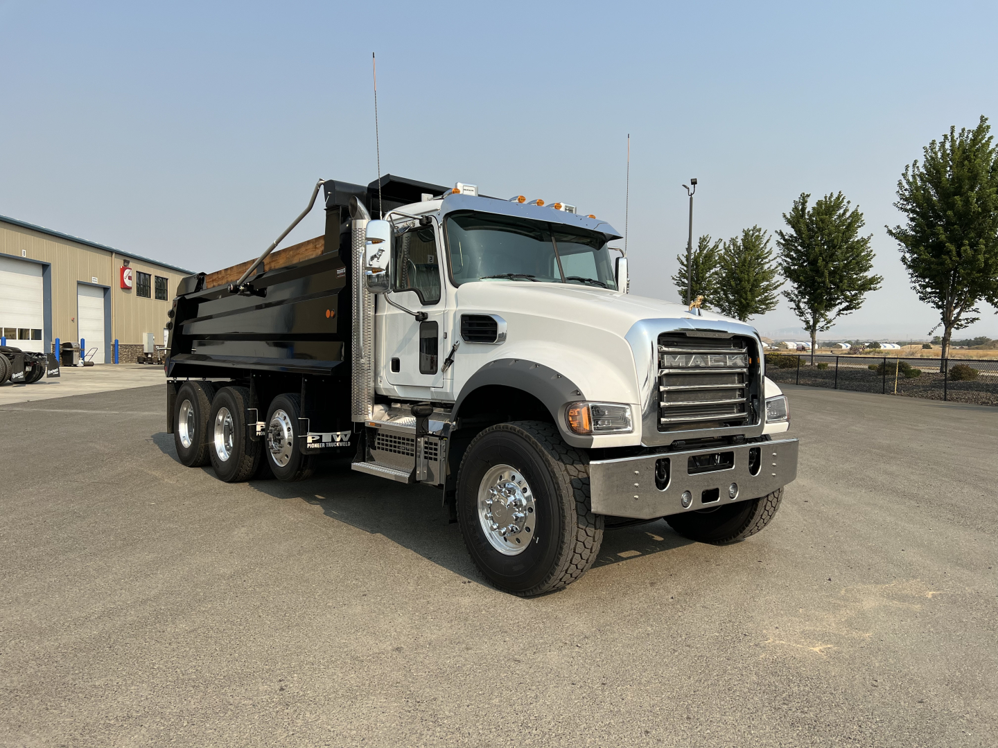 Dump truck with gray cab and black dump bed, front angled view, Mack grille, three-axle configuration, wheels and mirrors visible.