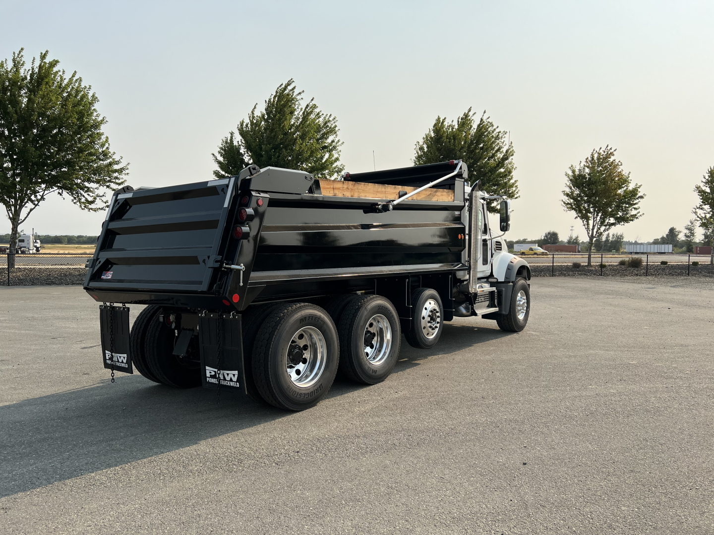 Dump truck rear and side view showing three-axle tandem drivetrain, black dump bed, heavy-duty tires, chrome wheels, and Mack branding on mud flaps.