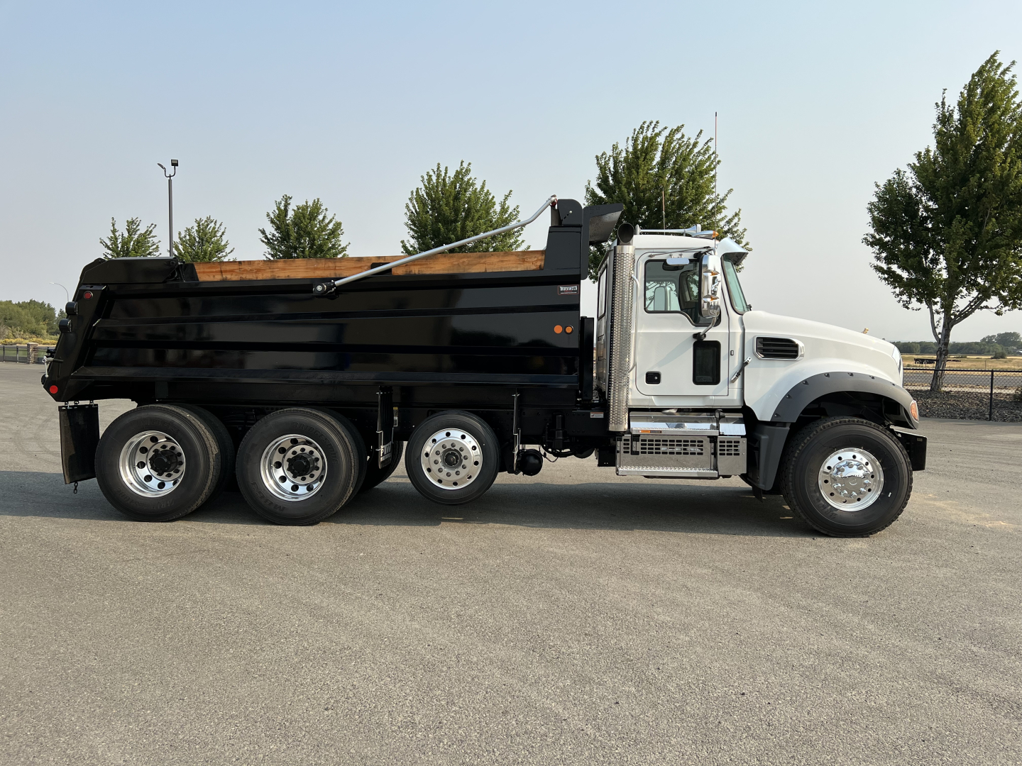 Dump truck side view showing Mack GR84F with black dump bed and white cab, six-wheel Rear, chrome steps, and long hydraulic arm on top.