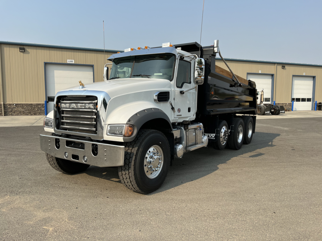 Dump truck front-left view showing Mack grille, chrome bumper, white cab, and black dump bed with hydraulic cylinders.