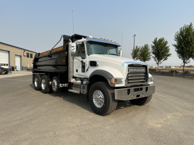 Dump truck with gray cab and black dump bed, front angled view, Mack grille, three-axle configuration, wheels and mirrors visible.