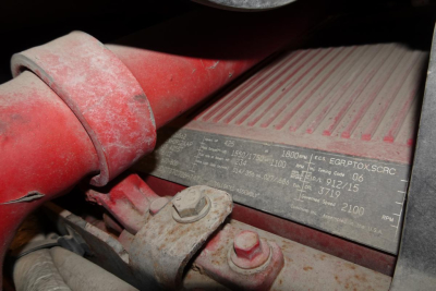 Label plate on engine bay showing specifications and serial numbers, attached to a red boot and metal bracket with a bolt.