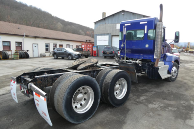 Kenworth T660 tractor chassis rear view showing dual rear axles and mud flaps with red reflectors. Blue cab with sleeper window, third axle not visible. Gravel lot and buildings in background.