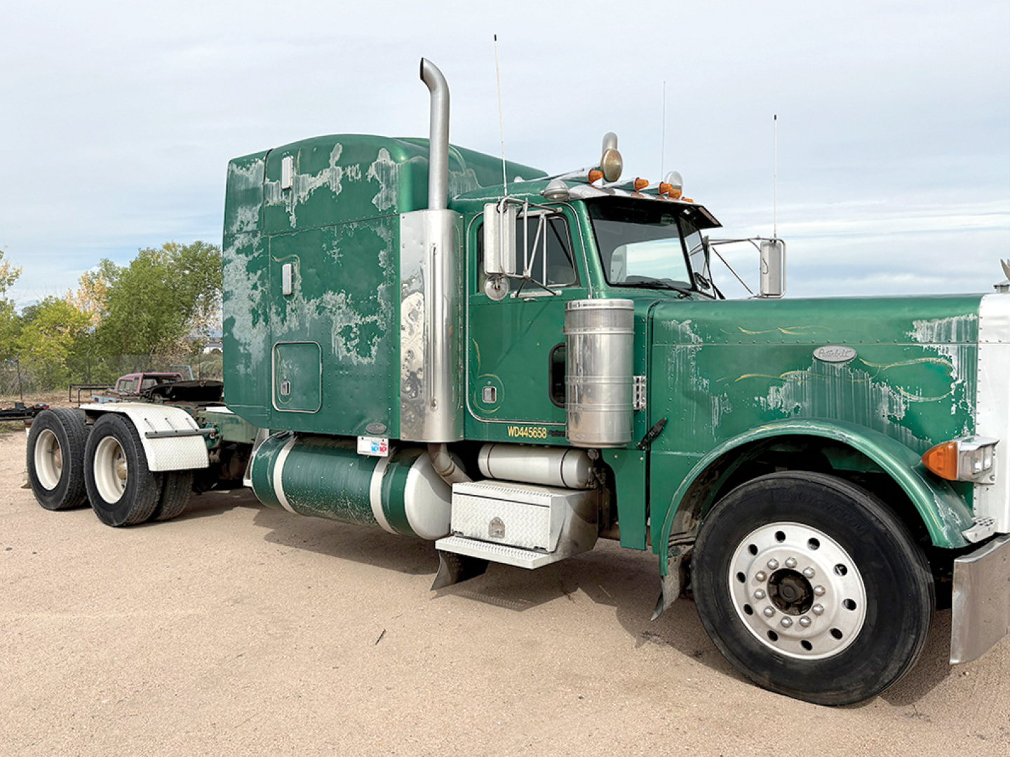 Peterbilt 379 conventional truck with extended hood, green paint, visible wear, side exhaust stacks and dual rear axles in a dirt lot.