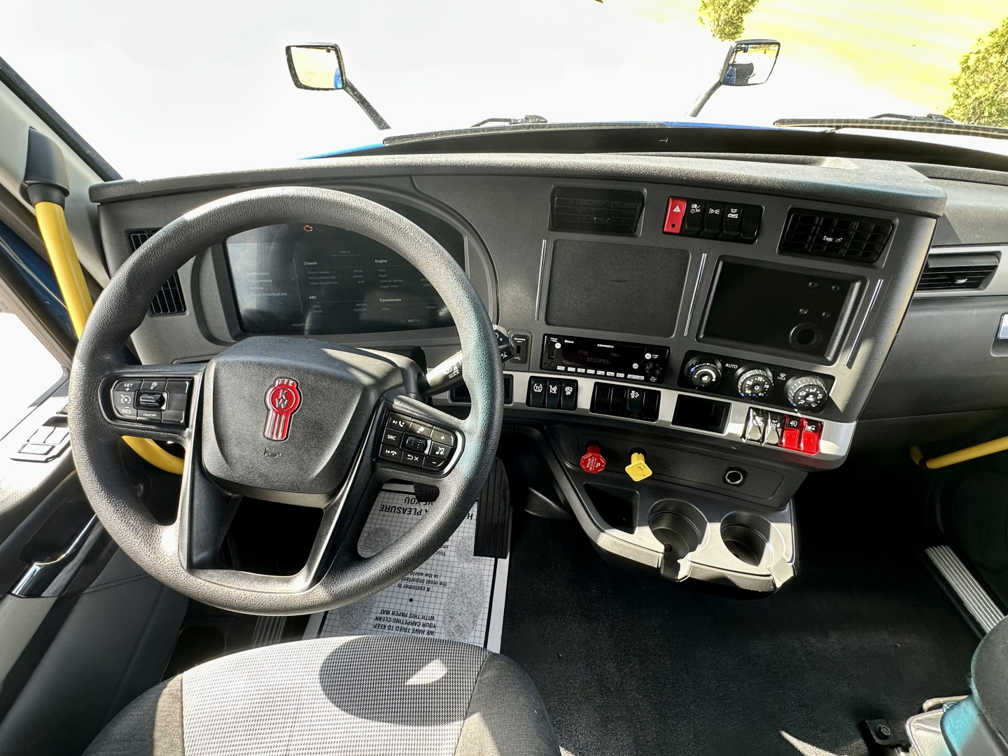 Dashboard and steering wheel of a 2023 Kenworth T680 cab interior with multiple gauges, a center touchscreen, and controls; visible label text on panels but no legible small print.