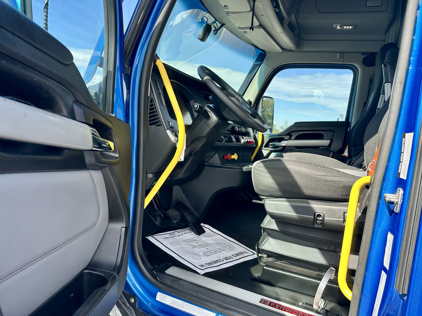 Kenworth T680 truck interior view showing driver's seat, steering wheel, dashboard, and entry door; yellow handrails and blue exterior visible.