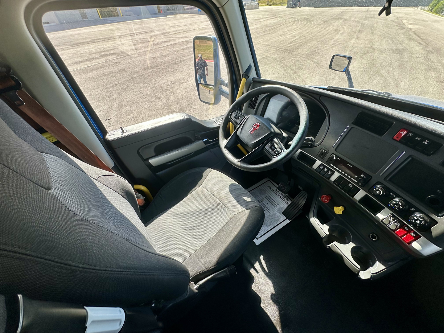 Kenworth T680 interior from driver side showing steering wheel, dashboard, pedals, and seating; side mirror visible; window view of open area.