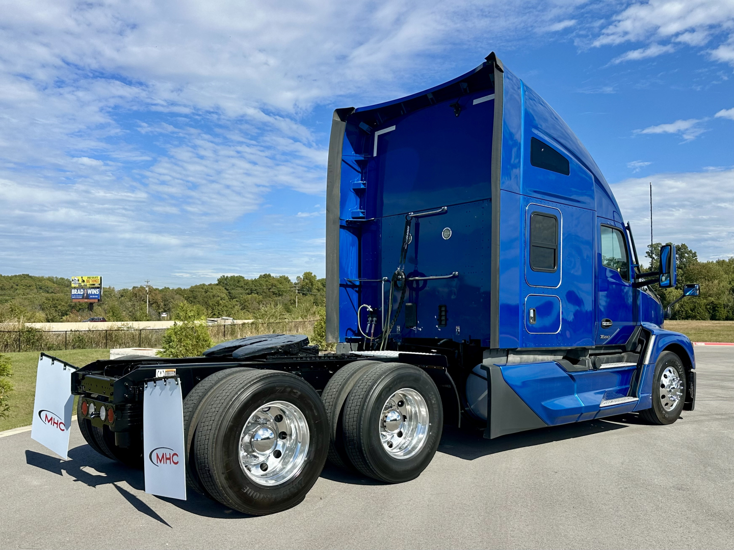 Blue 2023 Kenworth T680 semi tractor cab from rear three-quarter view showing triple rear axles, chrome fuel tanks, and rear fenders.